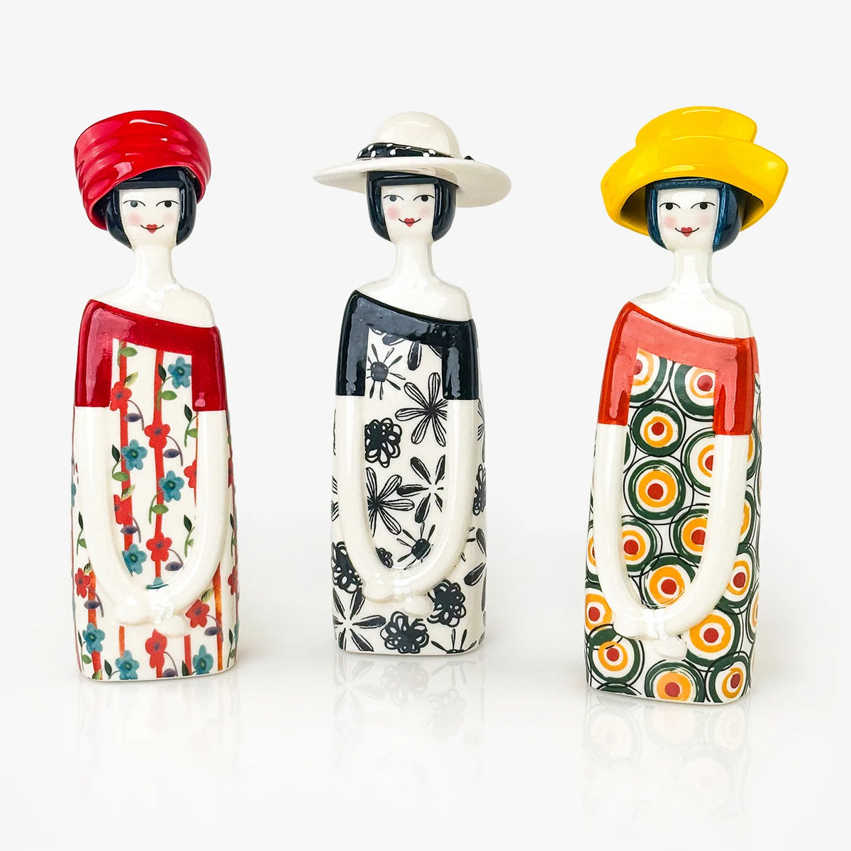 Three ceramic figurines standing side by side on a white reflective surface. The lady figurine on the left wears a red hat and a red-and-white patterned dress.