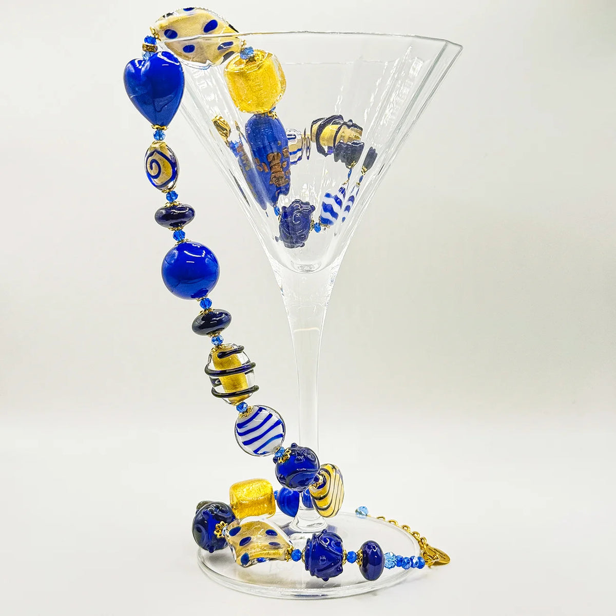 A glass beaded necklace with blue, yellow, and striped glass beads draped over a clear martini glass on a white background.