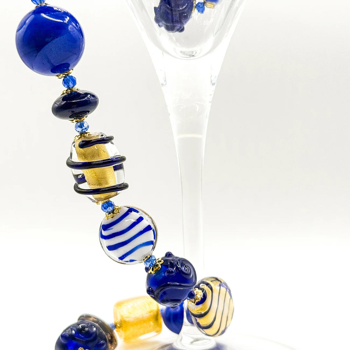 A close-up of a glass beaded garland or ornament threaded with glossy blue, white, and gold lampwork beads, shown draped over a clear glass stemware stem against a white studio background.