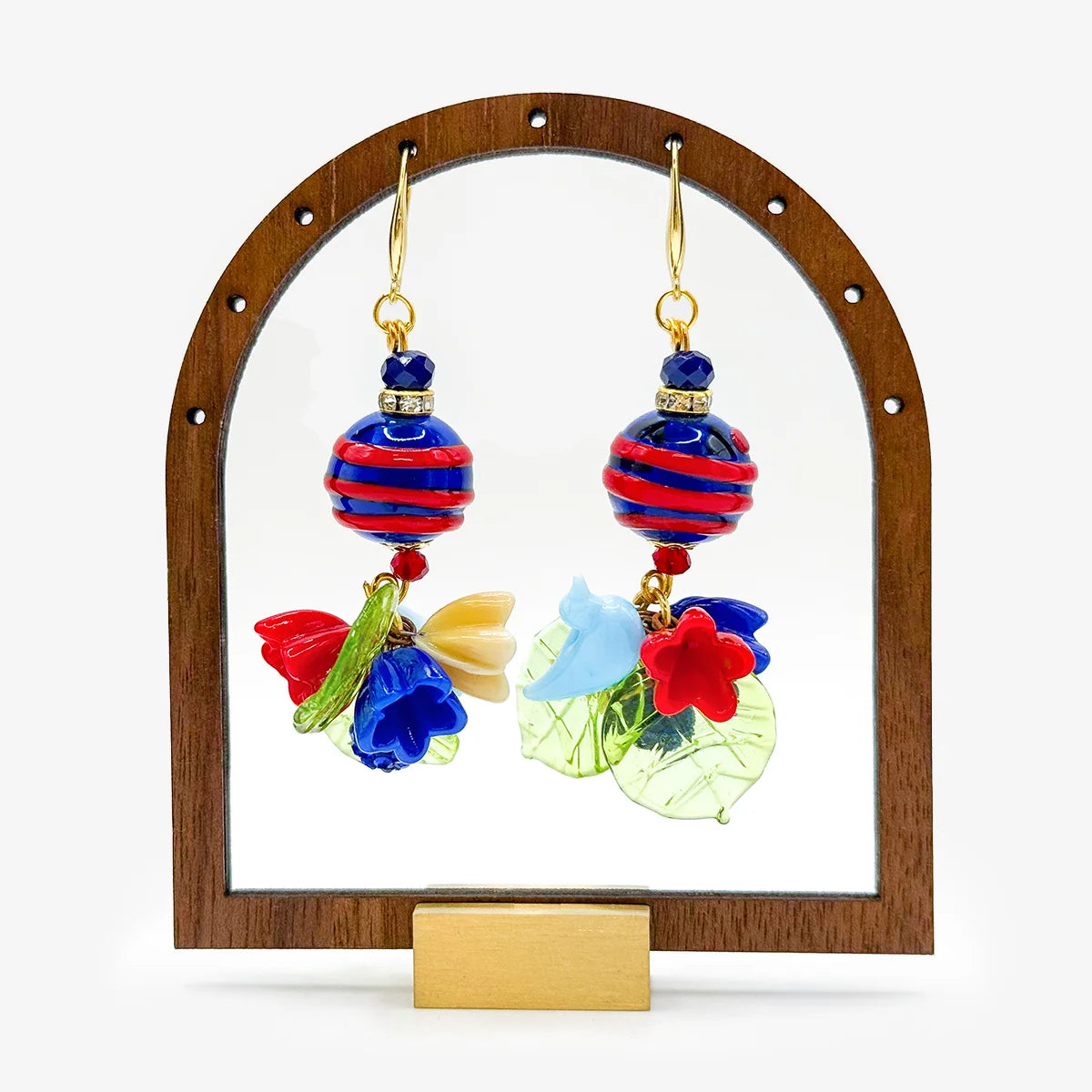 A jewelry set featuring a beaded bracelet and matching dangle earrings. The bracelet and earrings are decorated with red and black patterned beads, red and blue glass beads, a sheer red and blue ribbon bow, and colorful leaf and flower charms in green, blue, and red on gold-tone hooks.