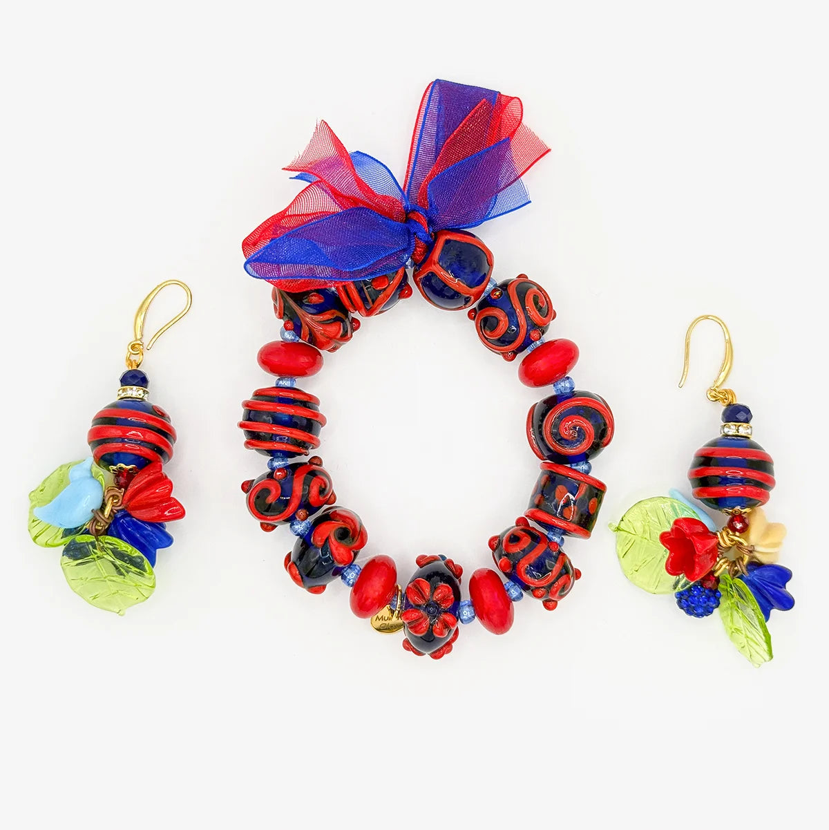A jewelry set featuring a beaded bracelet and matching dangle earrings. The bracelet and earrings are decorated with red and black patterned beads, red and blue glass beads, a sheer red and blue ribbon bow, and colorful leaf and flower charms in green, blue, and red on gold-tone hooks.