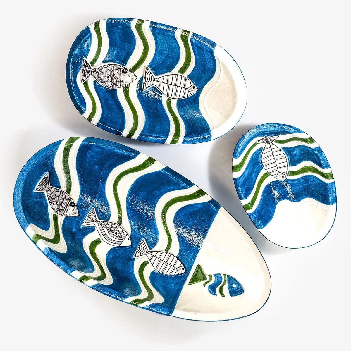 Three hand-painted ceramic fish-shaped plates in blue, green, and white, arranged on a clean white background.
