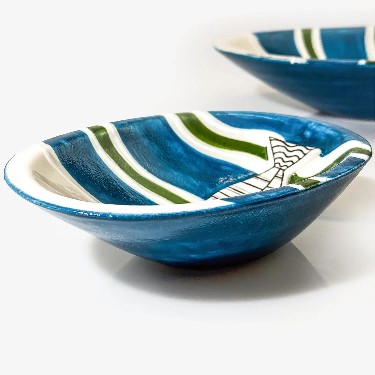 A close-up of a blue ceramic bowl with green and white stripes and a small white fish illustration, with a second matching bowl blurred in the background on a white surface.