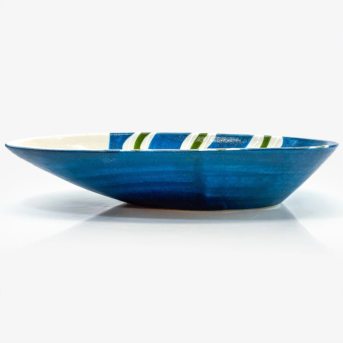 A handmade ceramic shallow bowl with a deep blue exterior, a cream-colored interior, and diagonal green and white stripes near the rim, shown on a white background.