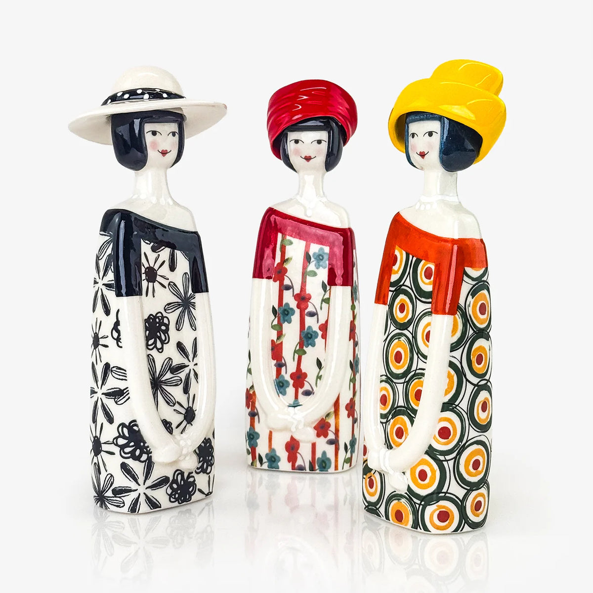 Three hand-painted ceramic dolls with stylized faces, standing side by side on a reflective white surface against a white background. The figurine on the left wears a white hat and a black-and-white patterned dress.