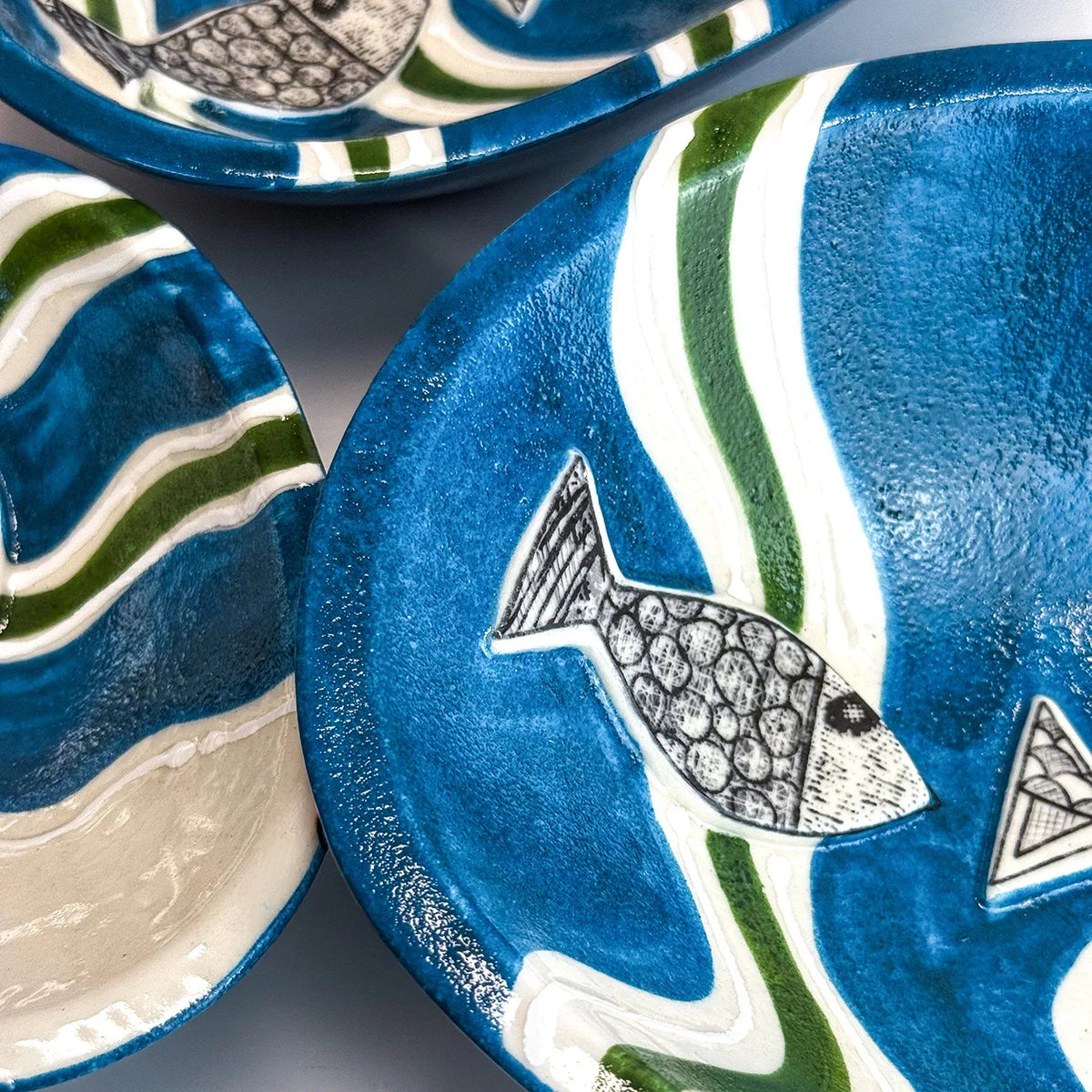 Close-up of ceramic plates with a blue and white wavy pattern, green accents, and fish motifs, shown on a neutral gray background.