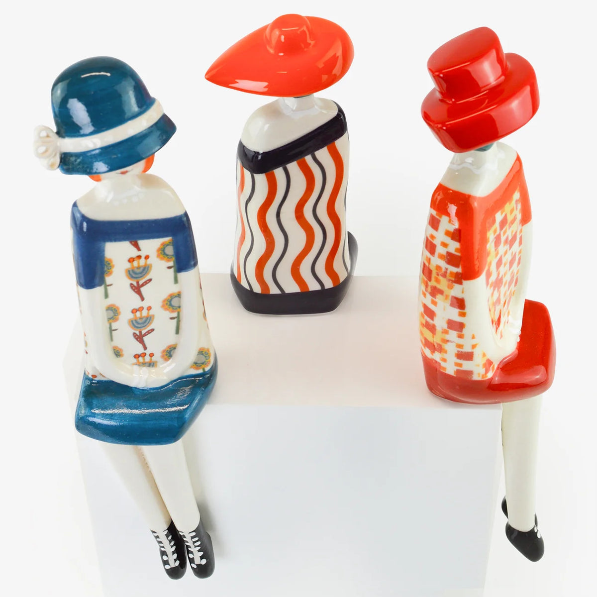 Three stylized ceramic figurines of seated people shown from the back, wearing colorful retro outfits and hats on a white background.