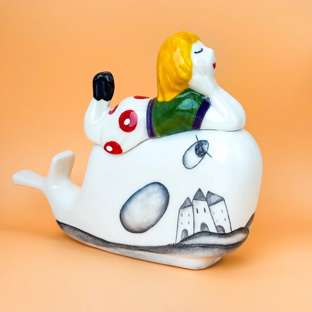 A whimsical ceramic figurine of a child with yellow hair reclining on a white whale decorated with a black-and-white castle scene. The figurine is photographed against a smooth peach-colored background.