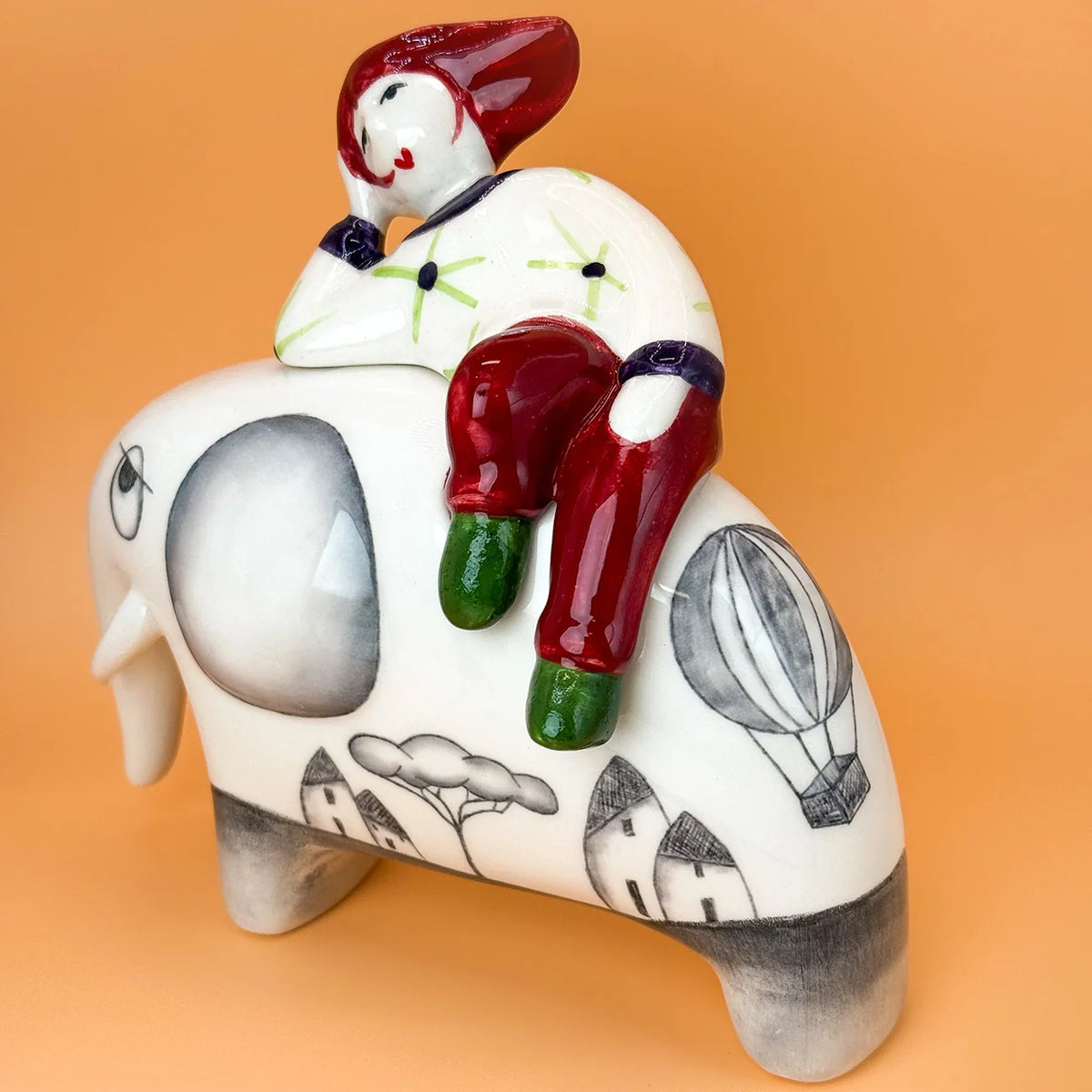A ceramic figurine of a white elephant decorated with black line drawings of houses, trees, with a child sitting on its back, set against a solid orange background.