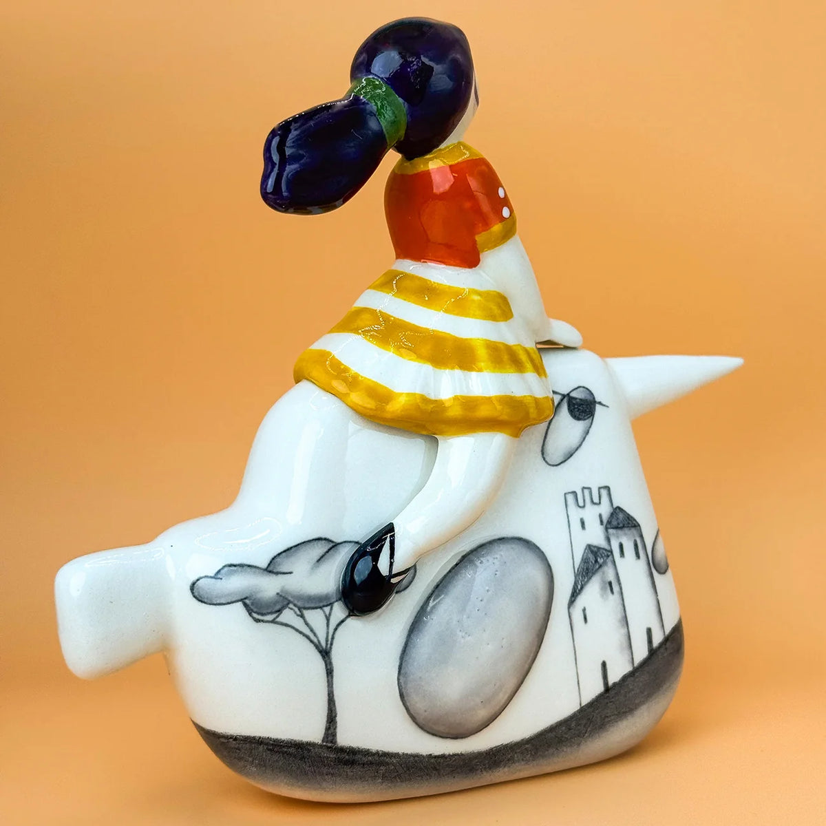 A hand-painted ceramic figurine of a child riding a narwhal. The narwhal is white with black and grey illustrations of a tree and a castle. the girl is wearing an orange and yellow dress, with black maryjane shoes, her hair is pulled back in a ponytail and appears to be flowing in the wind.