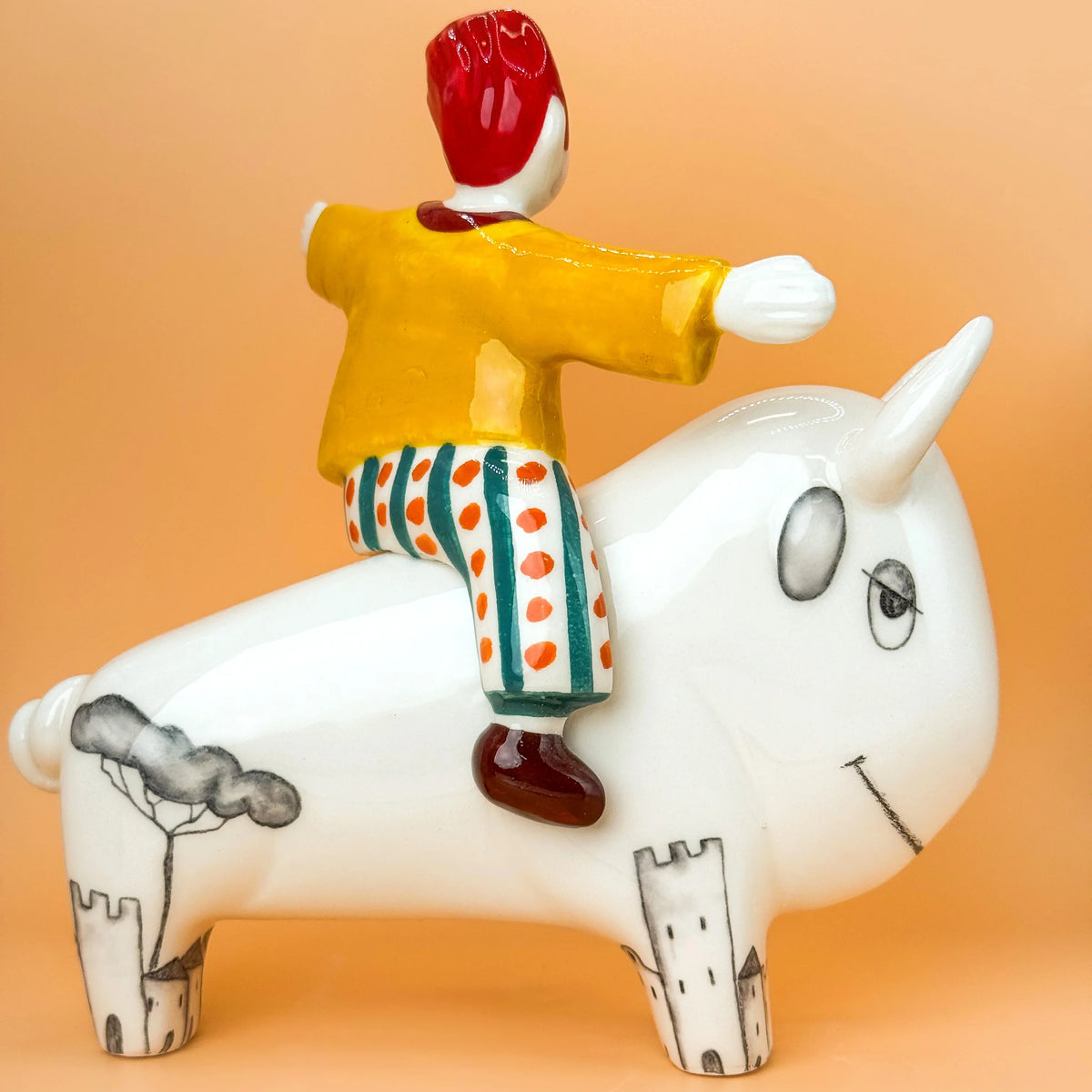 A ceramic figurine of a boy riding a white bison, set against a solid peach-colored background. The boy is wearing a yellow shirt, and green-and-white polka-dot pants, while the bull has hand-painted black details on its legs and a tree on its back.