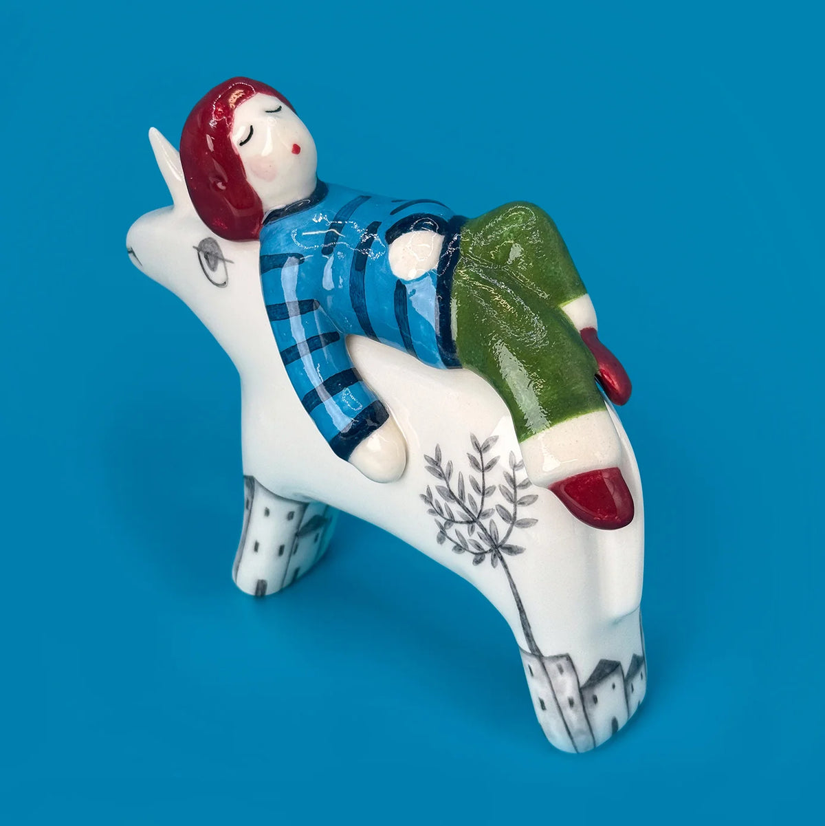 A ceramic figurine of a child with red hair lying on a white unicorn, set against a solid blue background. The figurine features a blue striped shirt, green pants, and red shoes, with black line drawings of a tree and houses on the unicorn's legs.