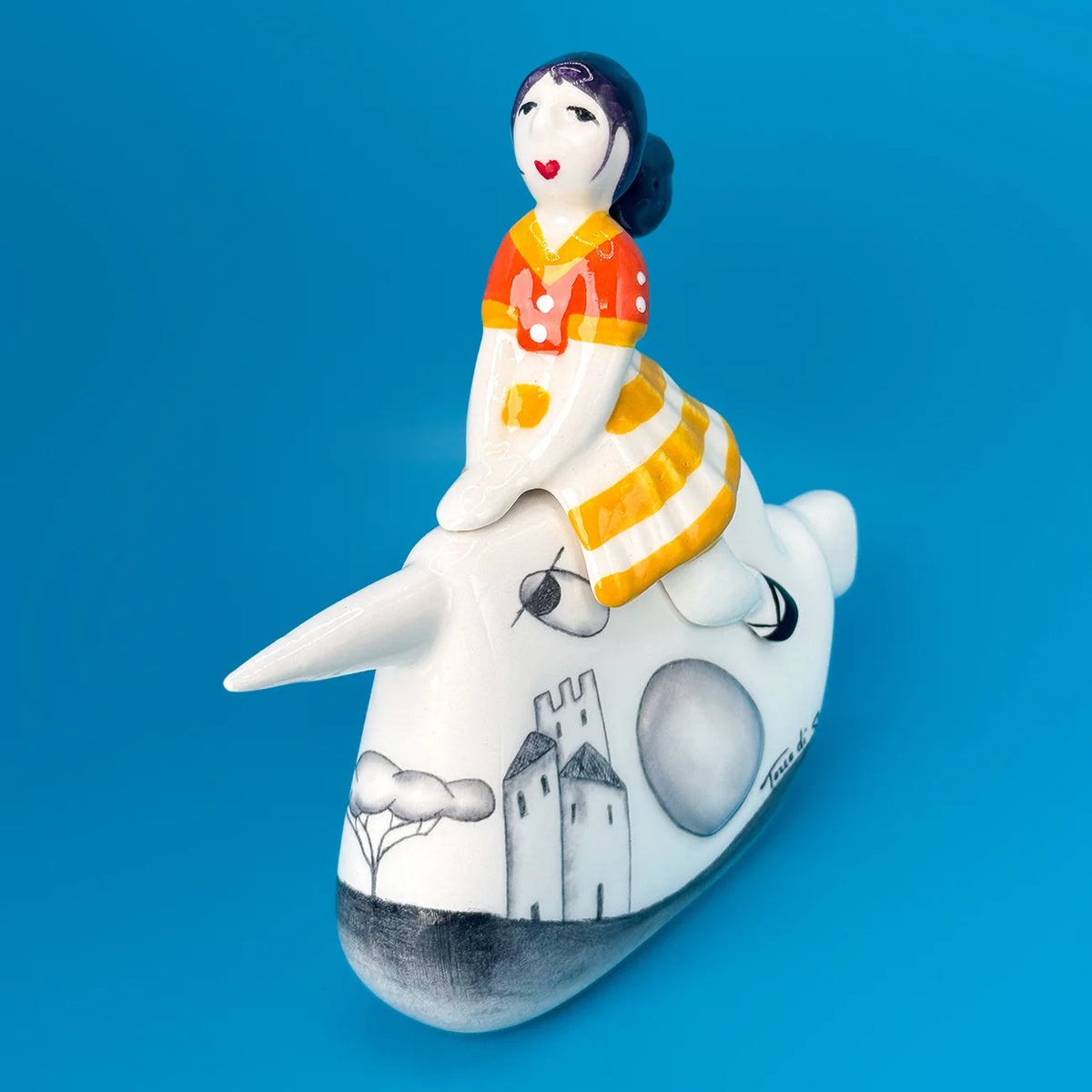 A hand-painted ceramic figurine of a girl riding a narwhal, set against a solid blue background. The narwhal is white with black hand-drawn landscape details, and the rider is dressed in a white, orange, and yellow dress.
