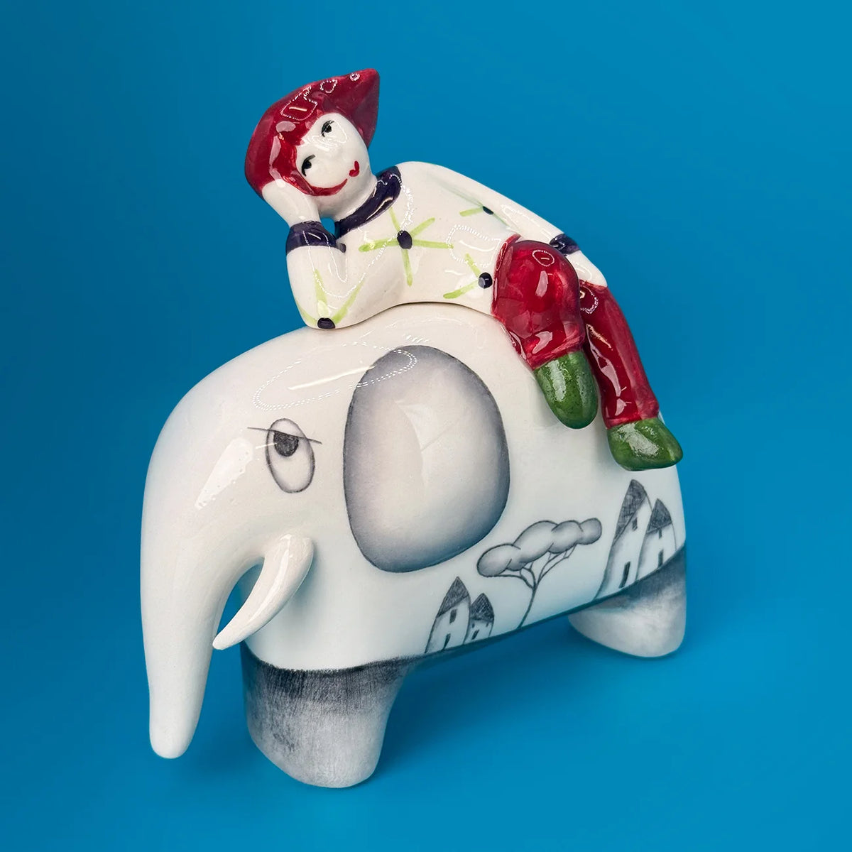 A ceramic figurine of a child riding an elephant, set against a solid blue background. The child is dressed in a white shirt with green leaf patterns, red pants, and has red hair, while the elephant is white with gray ear and eye details, black line drawings of houses and trees, and gray legs.