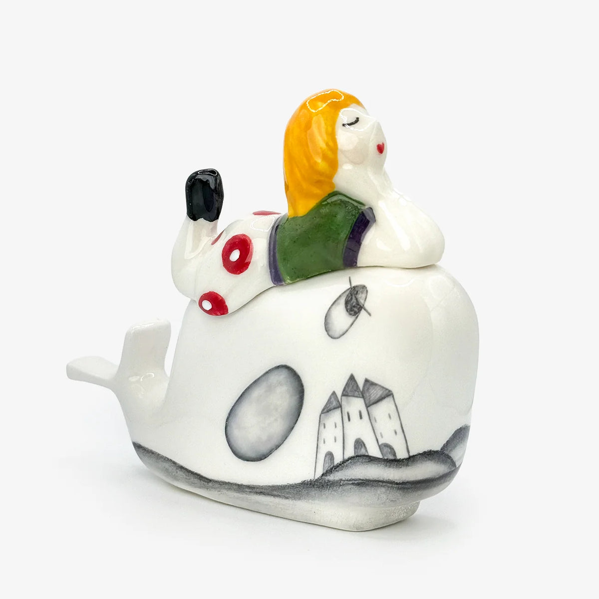 Ceramic figurine with a child laying on a whale. The whale is white with grey illustrations of houses on the body The child has a green shirt, polka dot pants, and yellow hair.