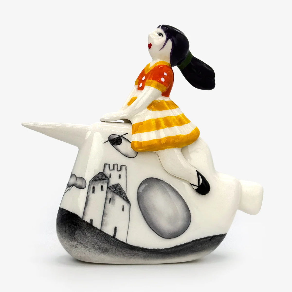A ceramic figurine of a girl riding a narwhal, set against a plain light gray background. The narwhal is white with black and gray painted details, and the rider wears a red and yellow striped dress with a black hair pony tail that appears to be flowing in the wind.