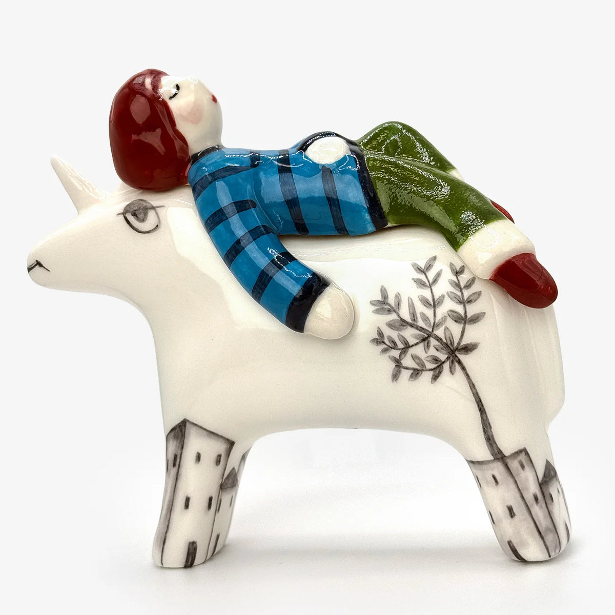 A ceramic figurine of a white ceramic animal with illustrations on legs and a tree painted on its side, with a small stylized person in a blue plaid shirt and green pants lying on its back, against a plain white background.