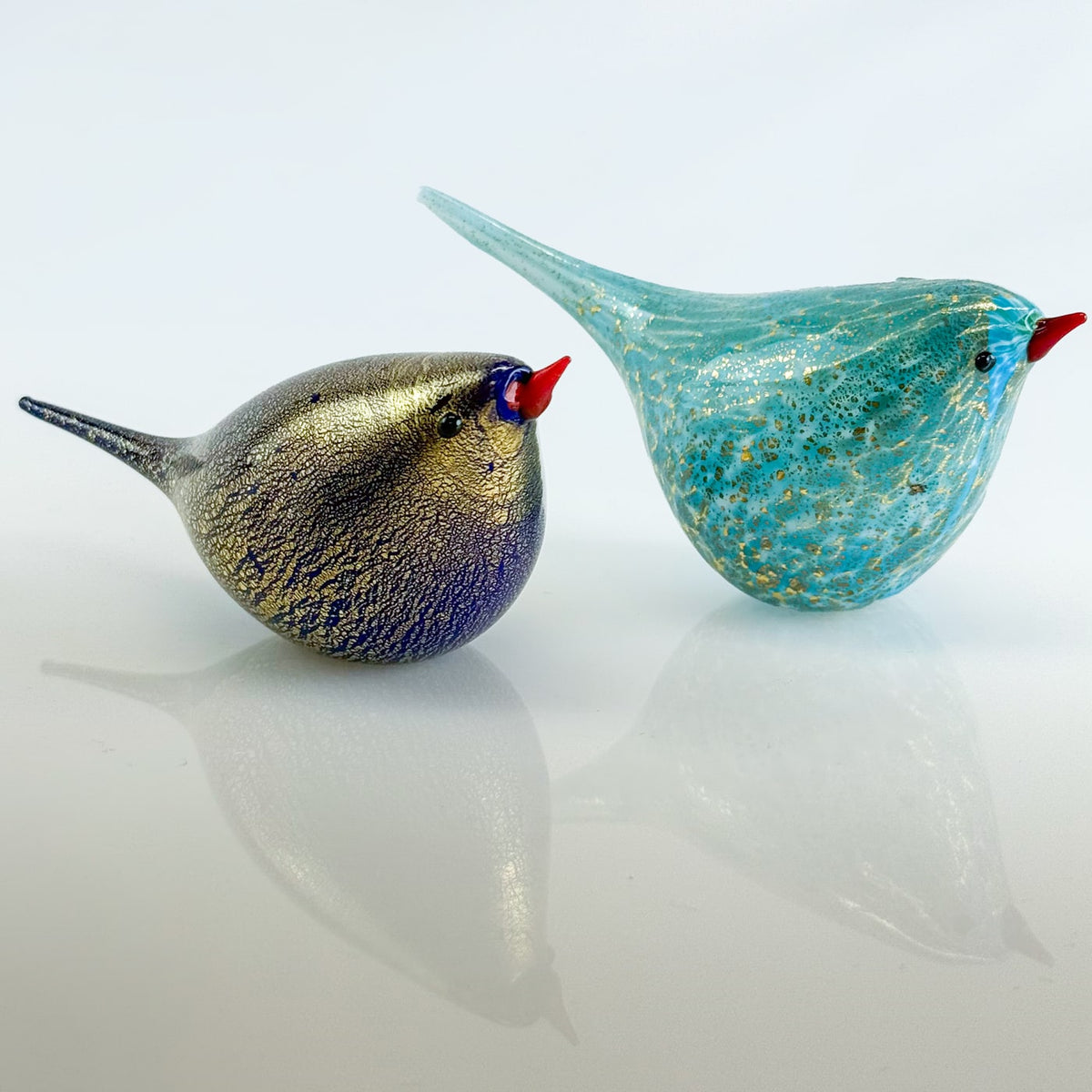 Two glass bird figurines, one gold and blue, the other turquoise with gold speckles, on a light gray background.