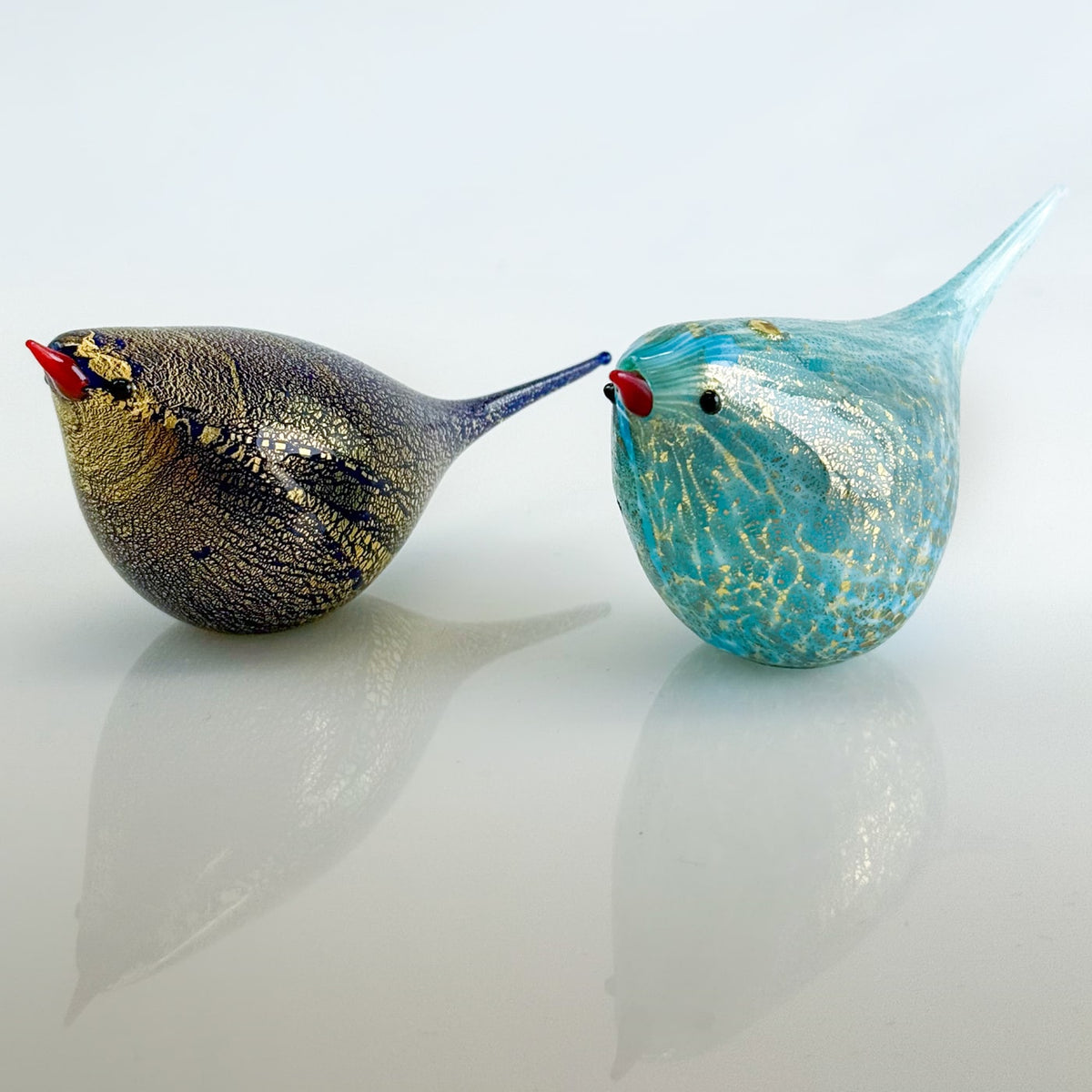 Two glass bird figurines, one gold and one blue with gold specks, on a reflective surface.