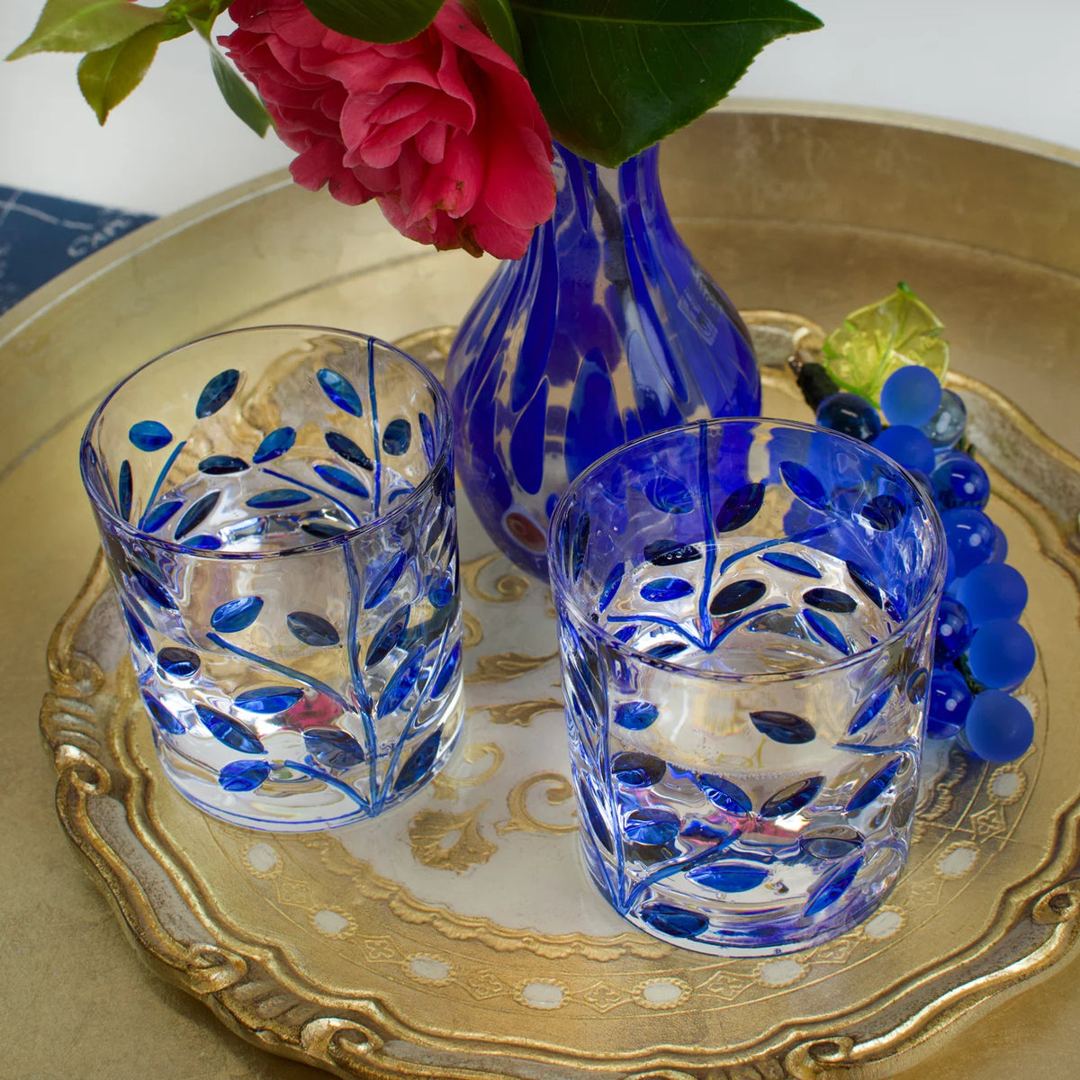 Two blue and clear decorated glasses with a blue Murano glass vase on a decorative Florentine wood tray.