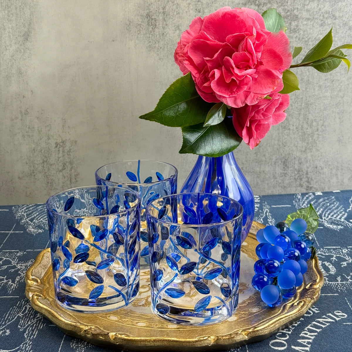 Set of blue glass tumblers with a blue vase and pink flowers on a gold tray.