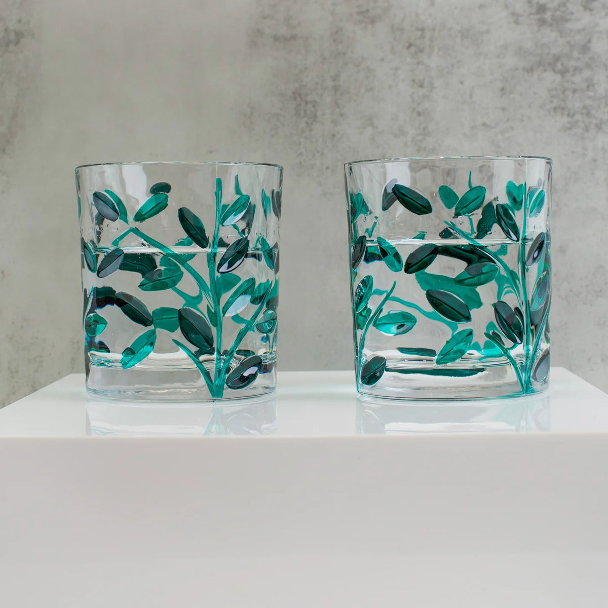 Two clear glasses with green leaf patterns on a white surface and gray background.