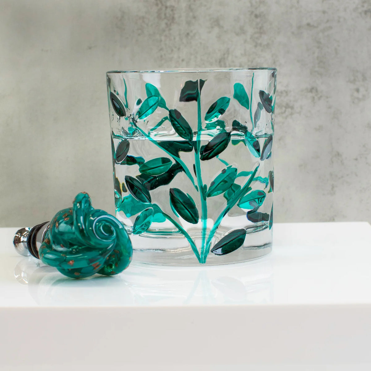 Clear glass tumbler with green rose wine stopper on a white surface and gray background.