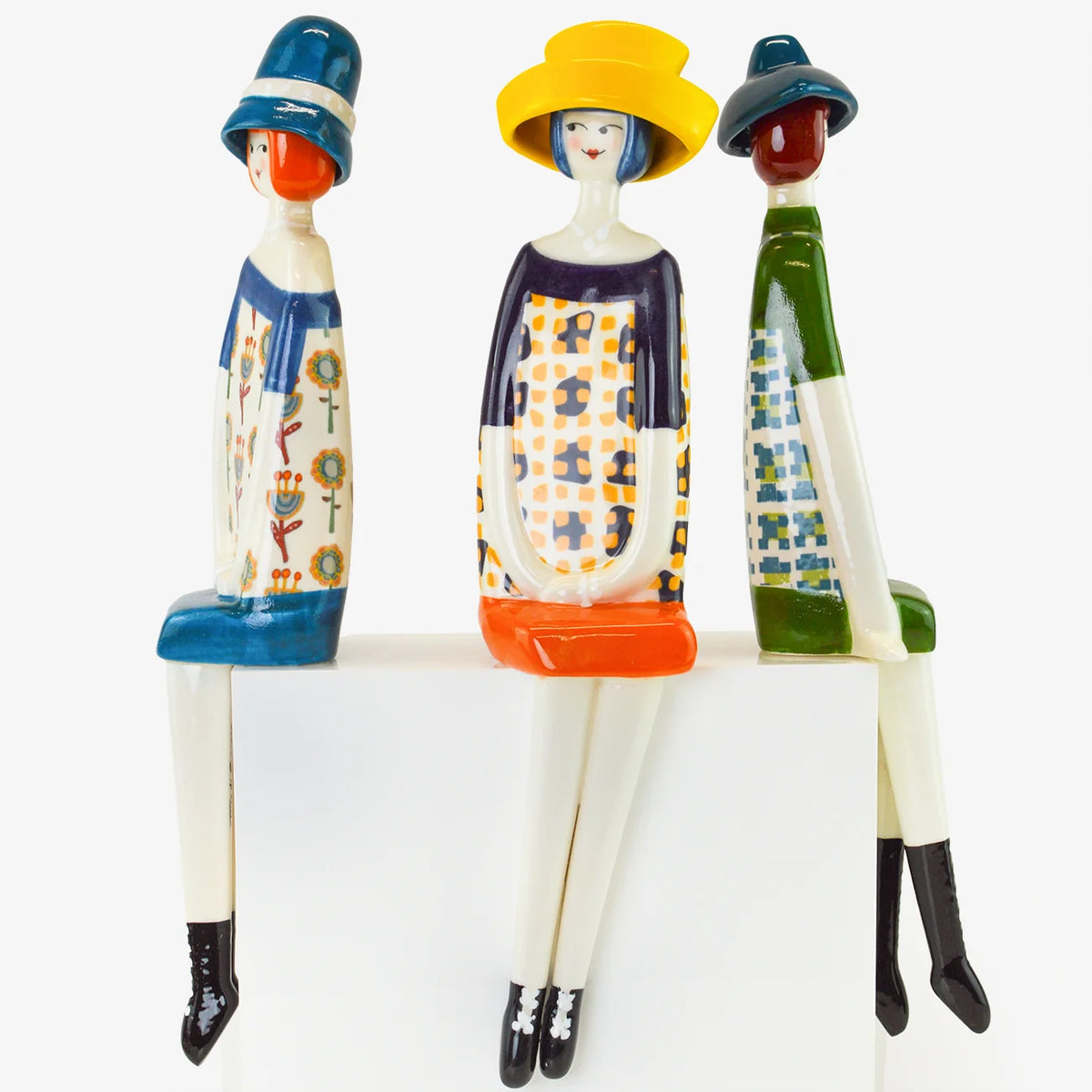 Three stylized ceramic figurines of seated women in colorful patterned clothing and hats, arranged side by side on a white surface against a light gray background.