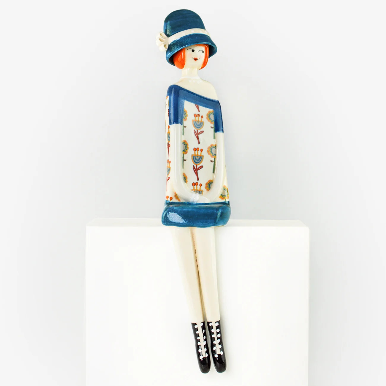 Three stylized ceramic figurines of seated women in colorful patterned clothing and hats, arranged side by side on a white surface against a light gray background.