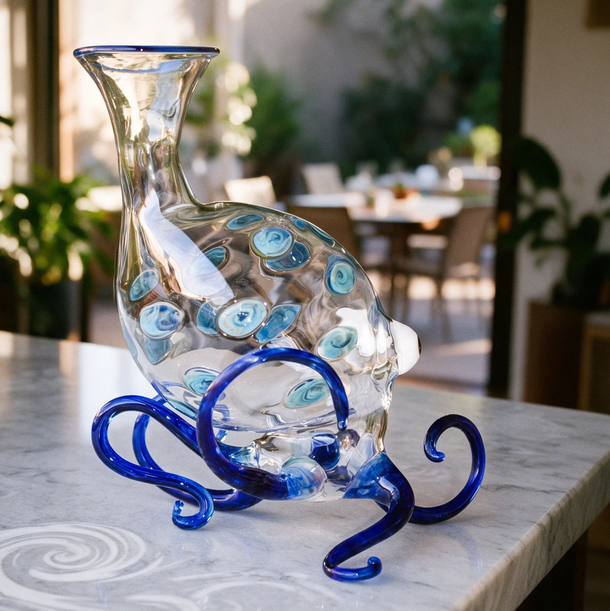 A clear glass decanter in the shape of an octopus with blue legs sits on a kitchen counter with a blurred dining room in the background
