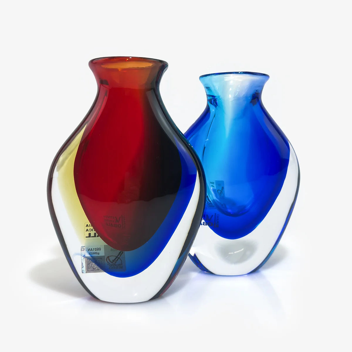 Two hand-blown glass vases with organic curved shapes, one featuring a gradient of red, dark brown, and blue over a clear base, and the other in a vibrant blue and white swirl pattern, set against a plain white background.