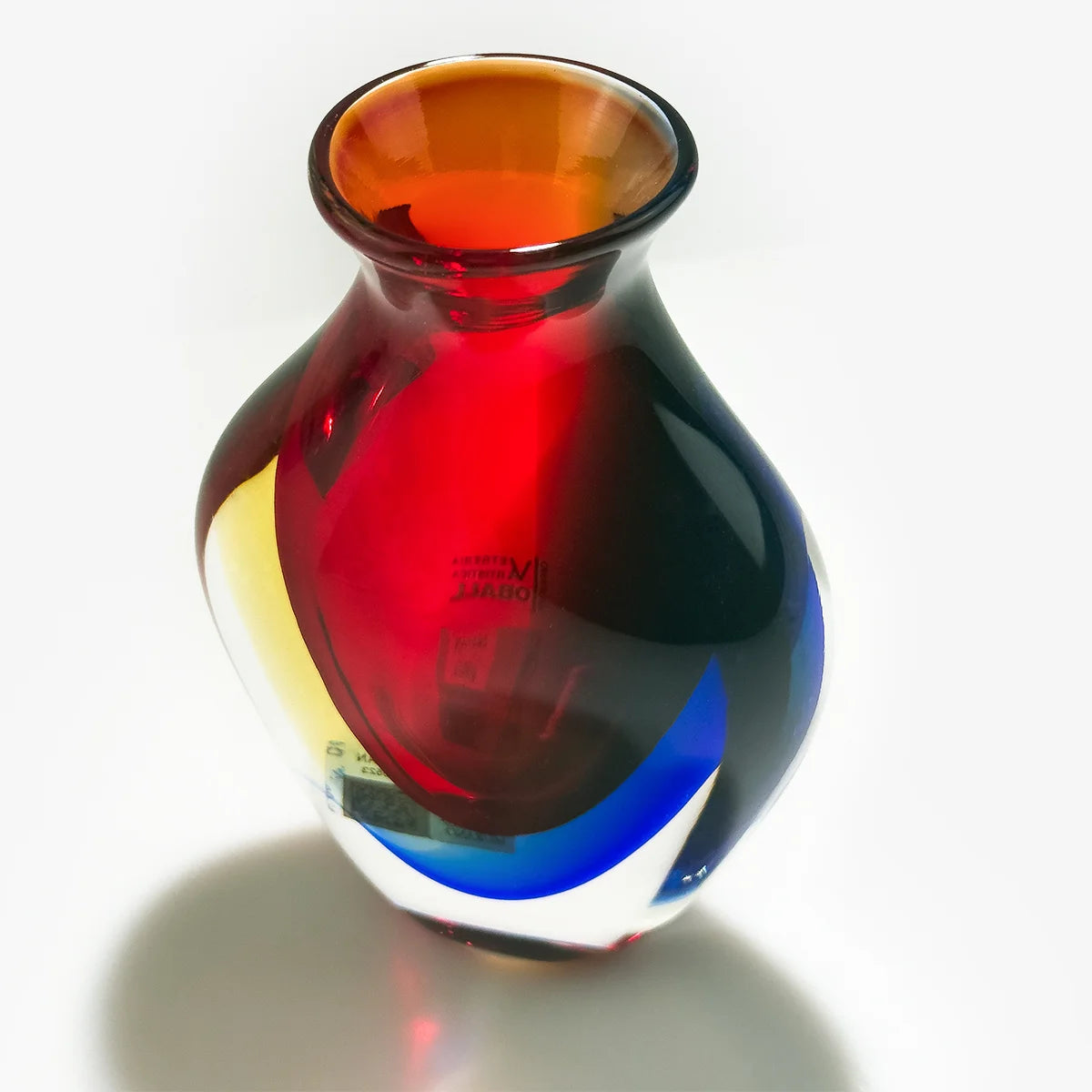 A hand-blown glass vase with a bulbous body, featuring a gradient of amber, translucent red, deep blue, and yellow on a white background.