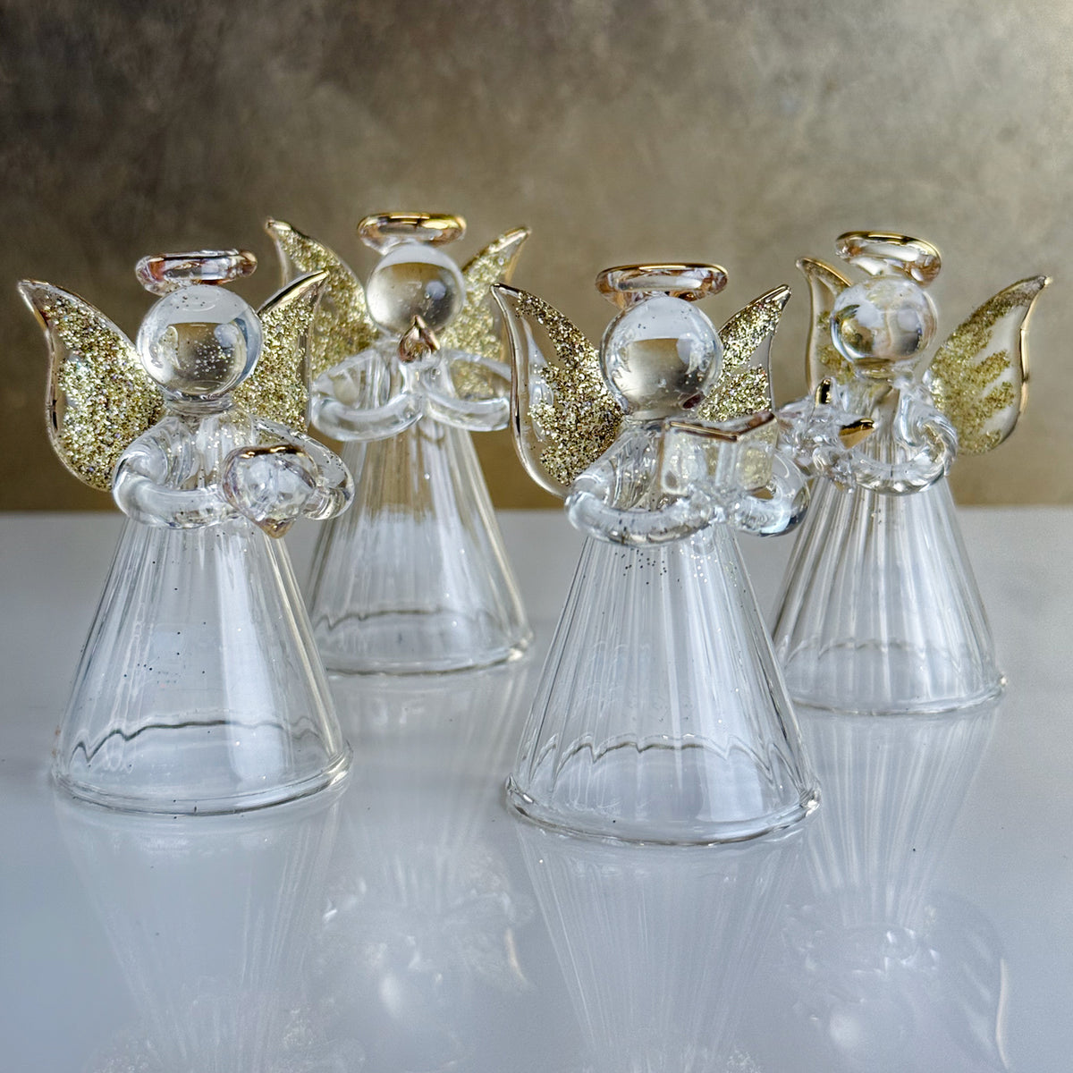 Four glass angel figurines with gold wings on a reflective surface.
