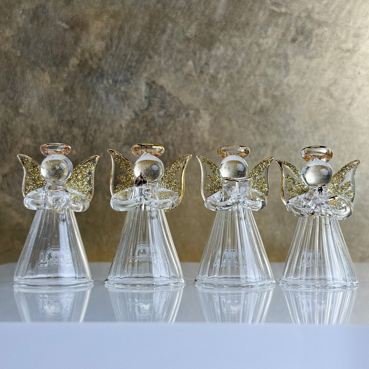 Four glass angel figurines with gold wings on a reflective surface with a textured brown background