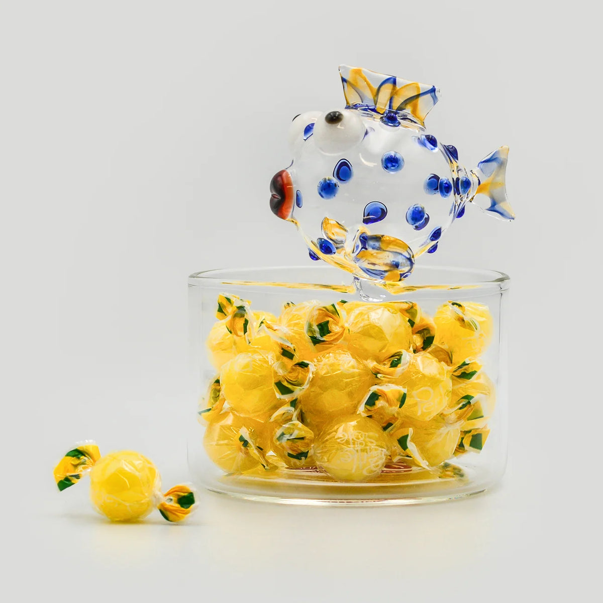 A clear glass with a decorative fish in blue, yellow, and white filled with individually wrapped hard lemon candies from Italy.