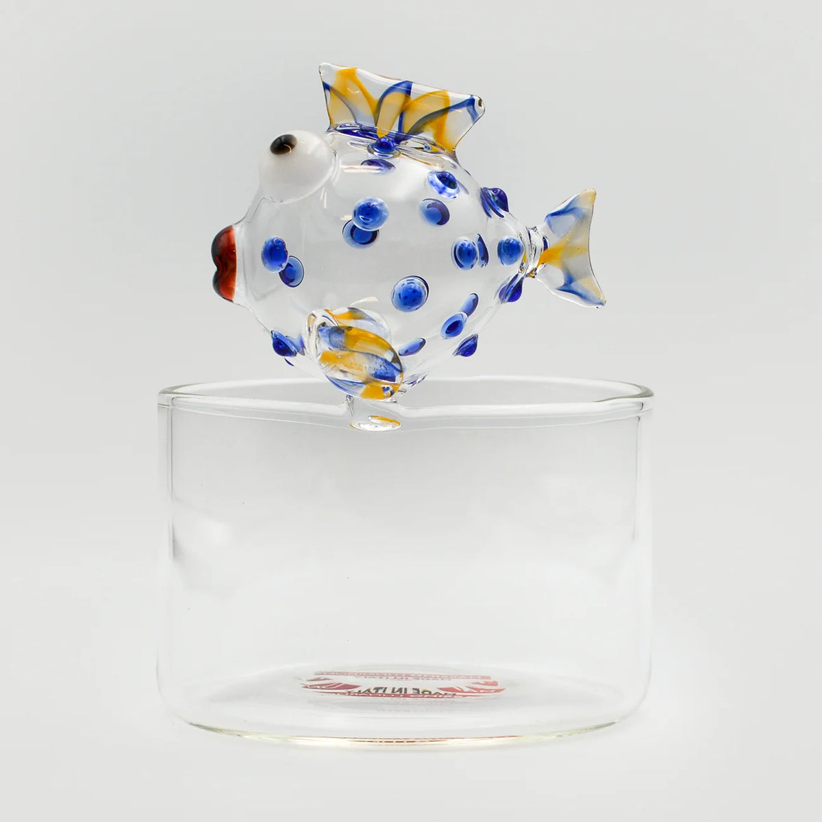 A hand-blown glass fish sculpture with blue and yellow details, perched on top of a clear small bowl against a light gray background.