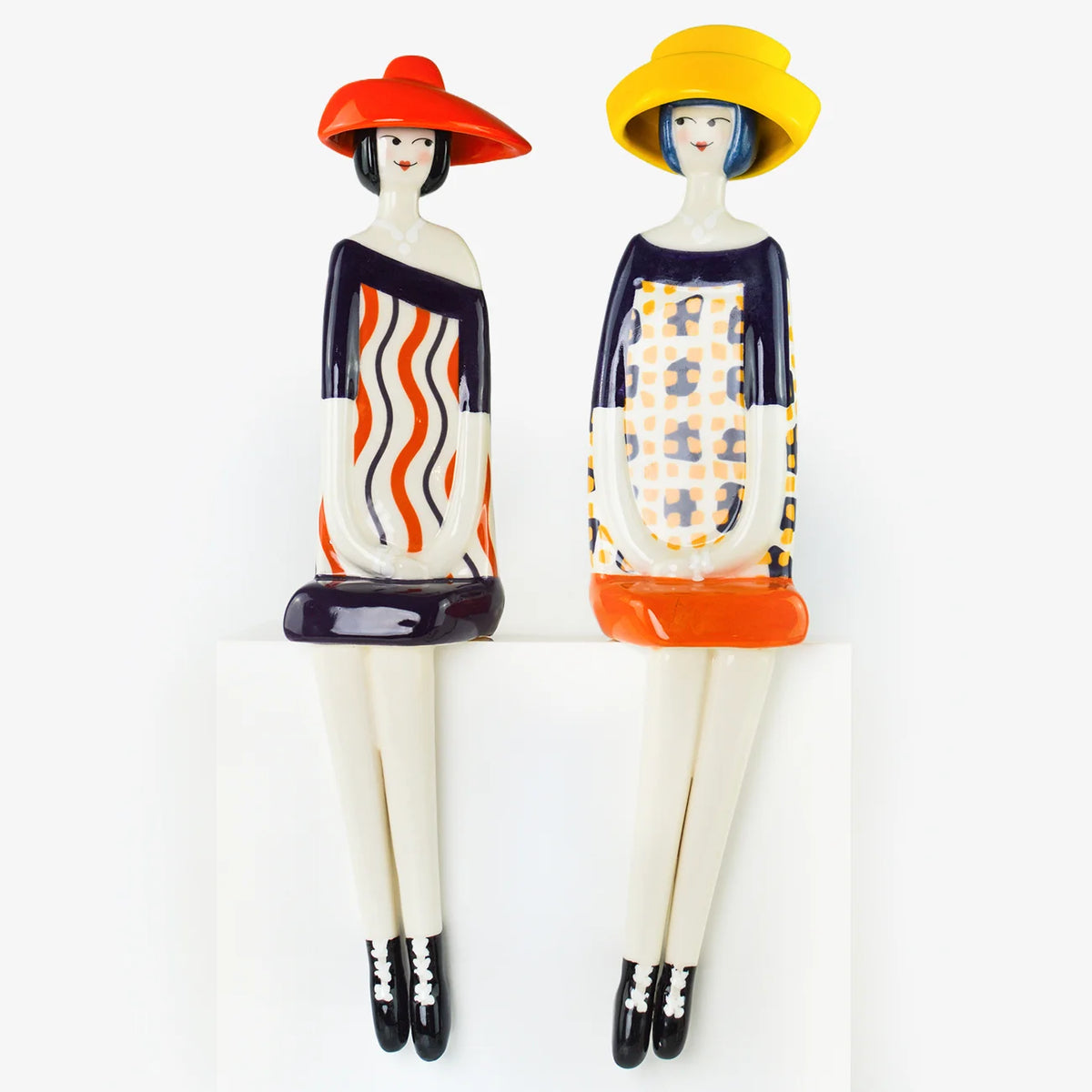 Two decorative figurines of stylized women seated side by side on a white block against a white background. One figurine wears a red hat and an off-white dress with orange and black wavy stripes; the other wears a yellow hat and a patterned off-white dress with orange and navy sections.