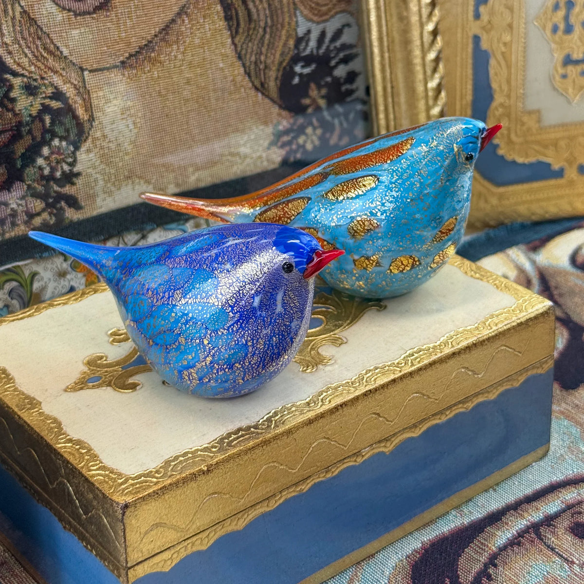 Colorful Murano glass Chirpie bird figurine in orange and teal, handblown by Rocco, displayed on a tapestry background.