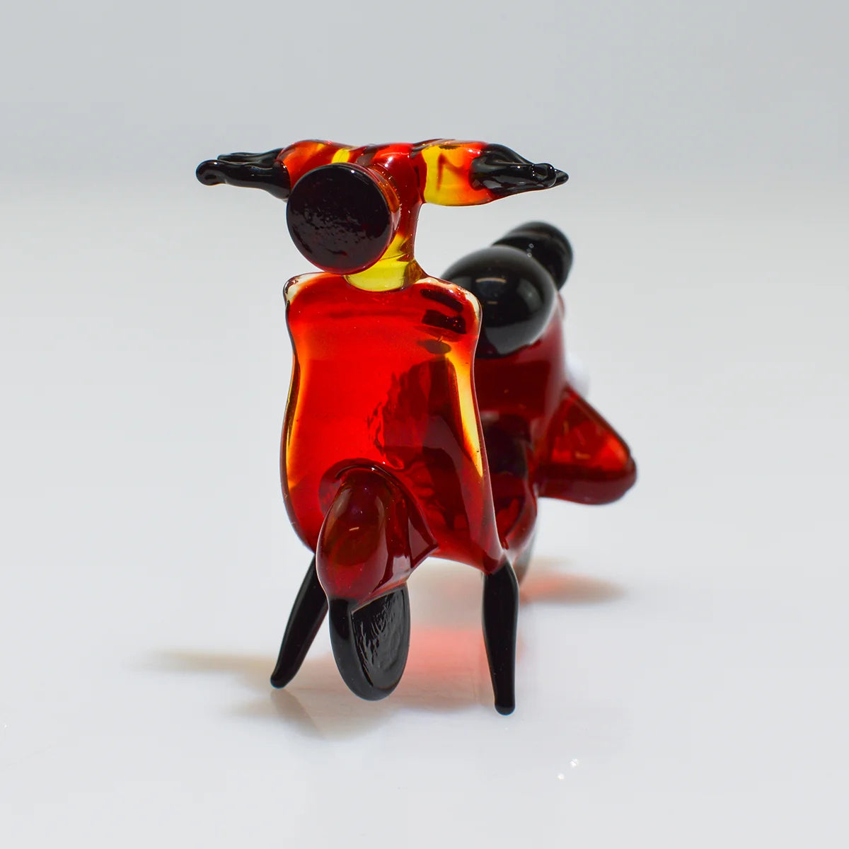 A small translucent glass sculpture of a scooter in glossy red with black and yellow accents, photographed on a clean white studio background.