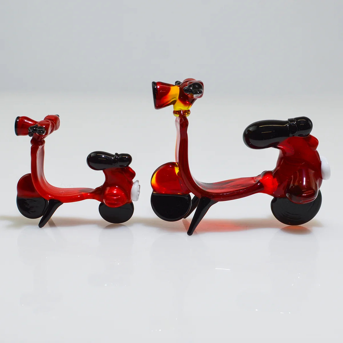 Two small red glass toy scooters with black wheels and seats, photographed on a clean white studio background.