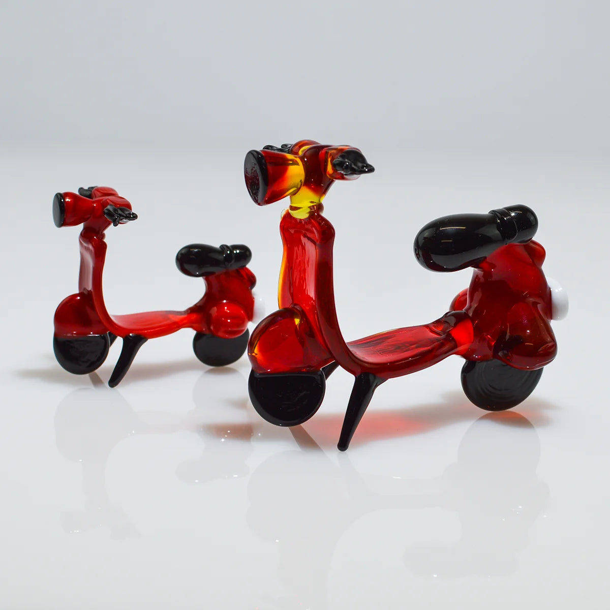 Two small translucent glass toy scooters on a white reflective surface against a light gray background. One scooter is made of bright blue glass, and the other is made of pale icy blue glass, both with dark purple seats and black wheels.