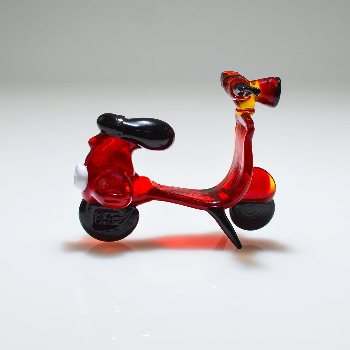 A small transparent glass toy scooter in red with black wheels, seat, and handlebars, photographed on a clean white studio background.
