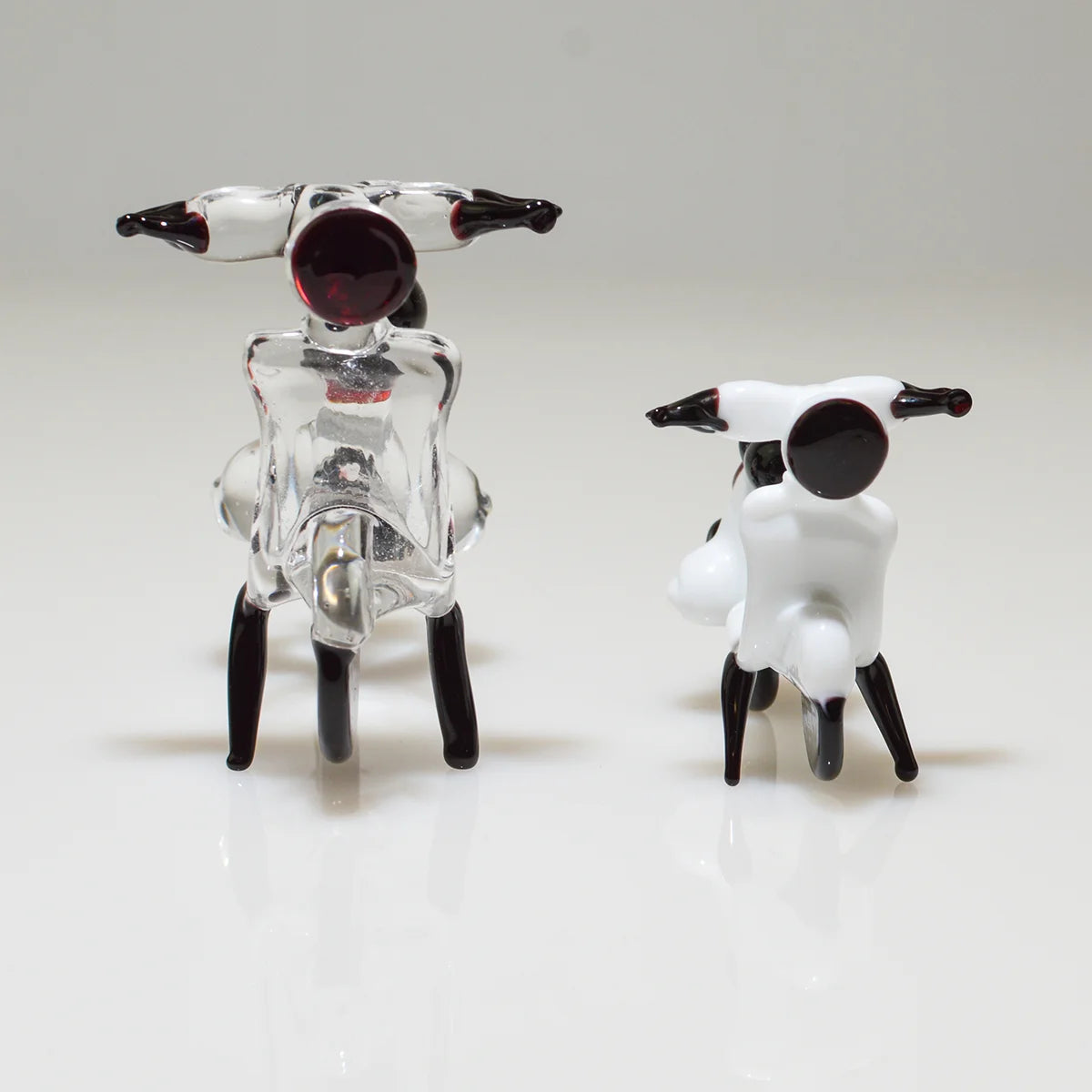 Two small decorative figurines on a white reflective surface against a light gray studio background. One figurine is transparent clear with black and dark red details, and the other is white with black details.