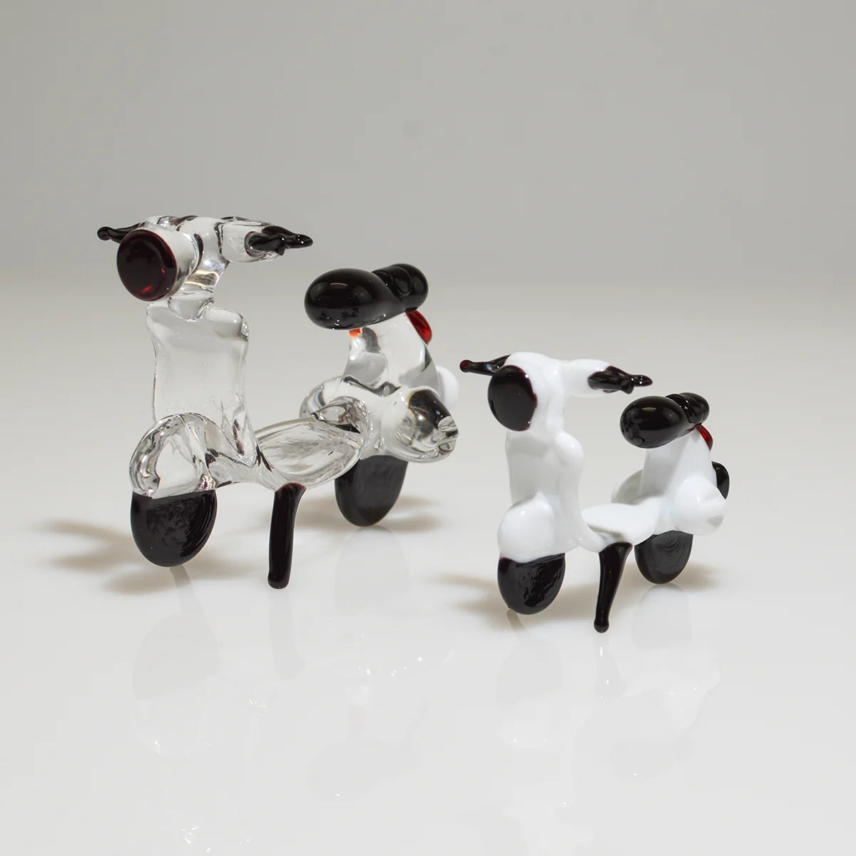 Three small glass scooter figurines in different sizes, featuring clear glass bodies with black wheels, seats, and handlebars, and small red neck details, arranged on a white reflective surface against a light gray background.