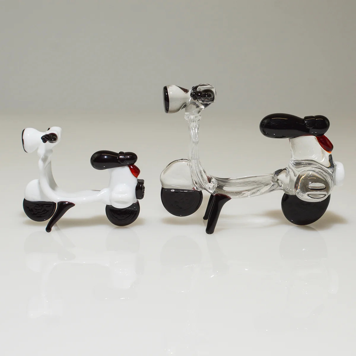 Two decorative glass toy scooters on a clean white reflective surface against a light gray background. One scooter is opaque white with black wheels, seat, and handlebars, and a small red accent.