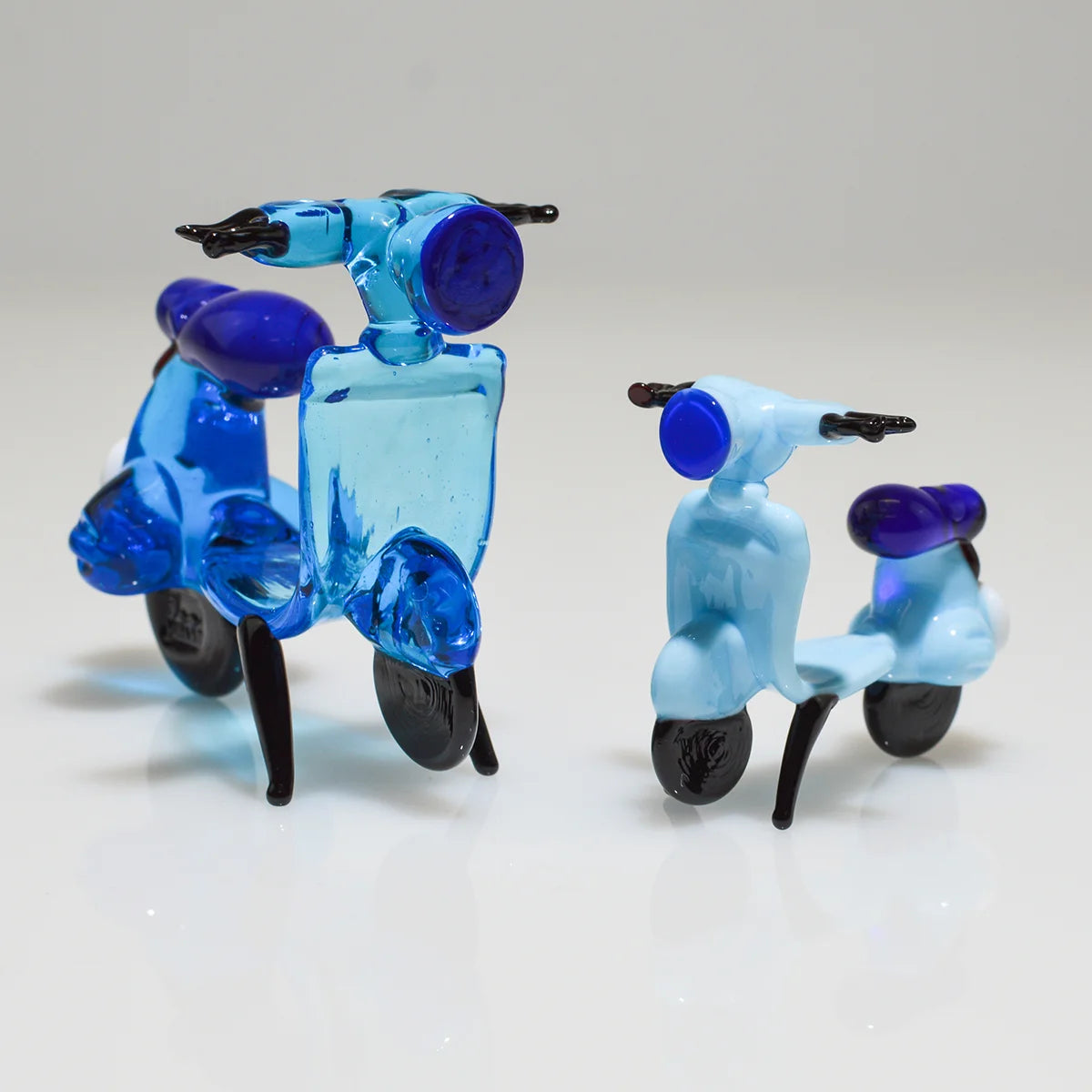 Two small translucent glass toy scooters on a white reflective surface against a light gray background. One scooter is made of bright blue glass, and the other is made of pale icy blue glass, both with dark purple seats and black wheels.