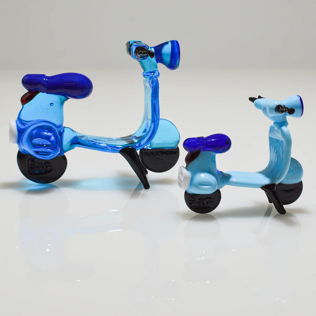 Two small translucent glass toy scooters, one larger in medium blue and one smaller in light blue, both with dark blue seats and black wheels, displayed on a white reflective surface against a light gray background.