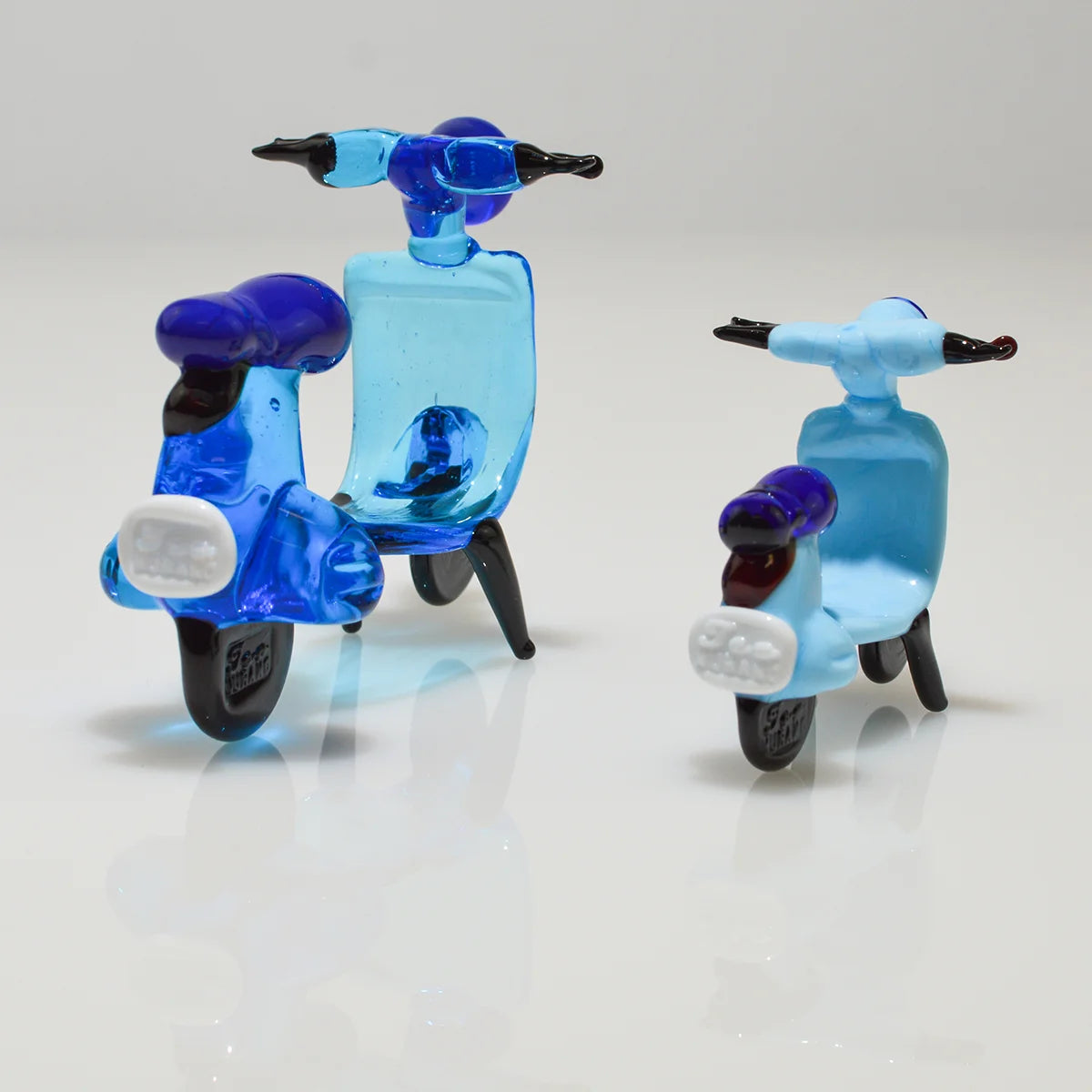 Two small translucent glass toy scooters on a clean white reflective surface. One scooter is a vibrant blue, and the other is a pale sky blue, both with dark purple seats and black handlebars.