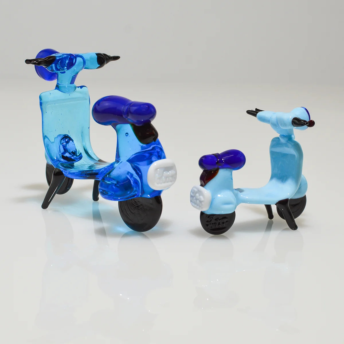 Two small glass toy scooters on a clean white reflective surface against a light gray studio background. One scooter is translucent aqua blue with dark blue seat and black wheels, and the other is pale blue with a dark blue seat and black wheels.