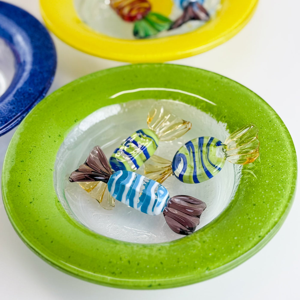 Colorful glass candy figurines on a green wide shallow bowl with other colored bowls in the background.